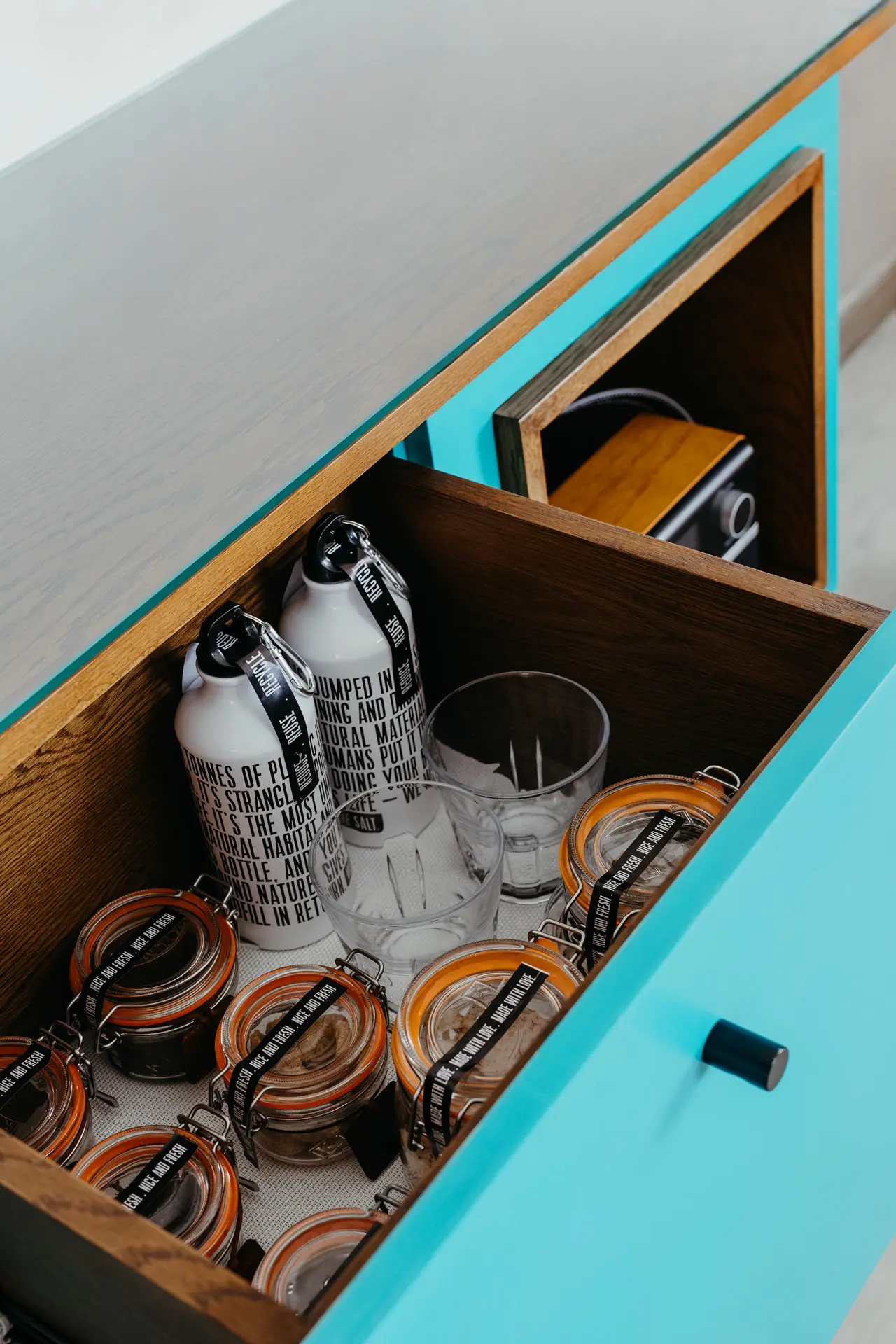 Hotel minibar drawer stocked with reusable water bottles, glassware and locally inspired refreshments.