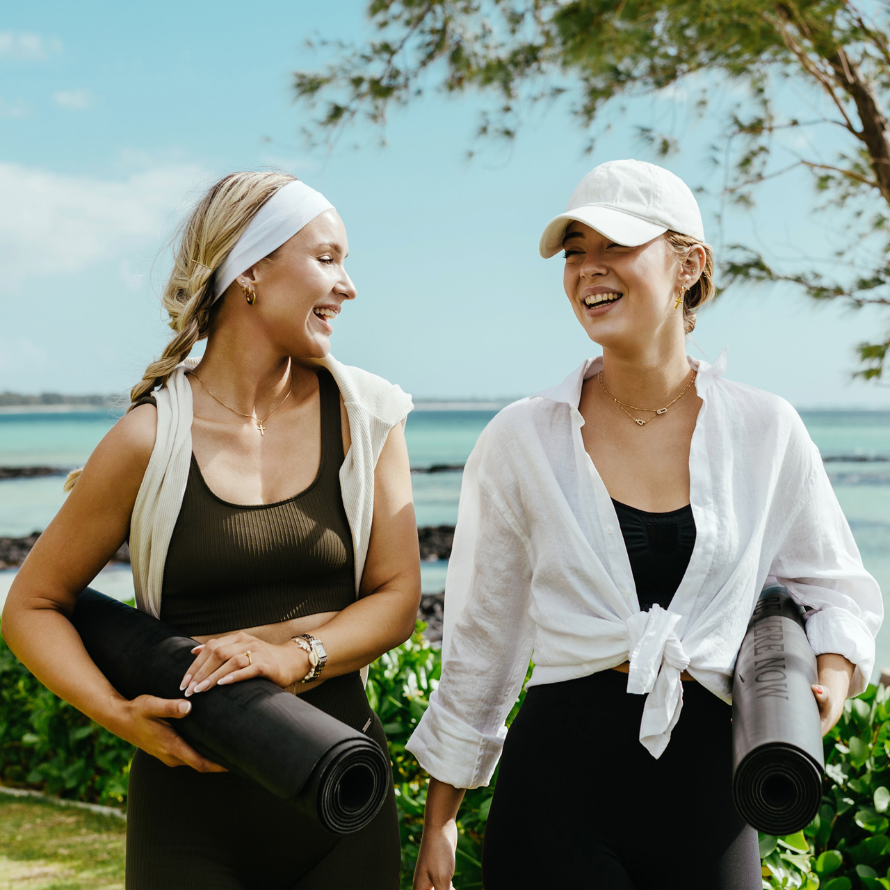Two guests carrying yoga mats stroll along the tropical coastline, enjoying a mindful wellness experience surrounded by nature and ocean views.