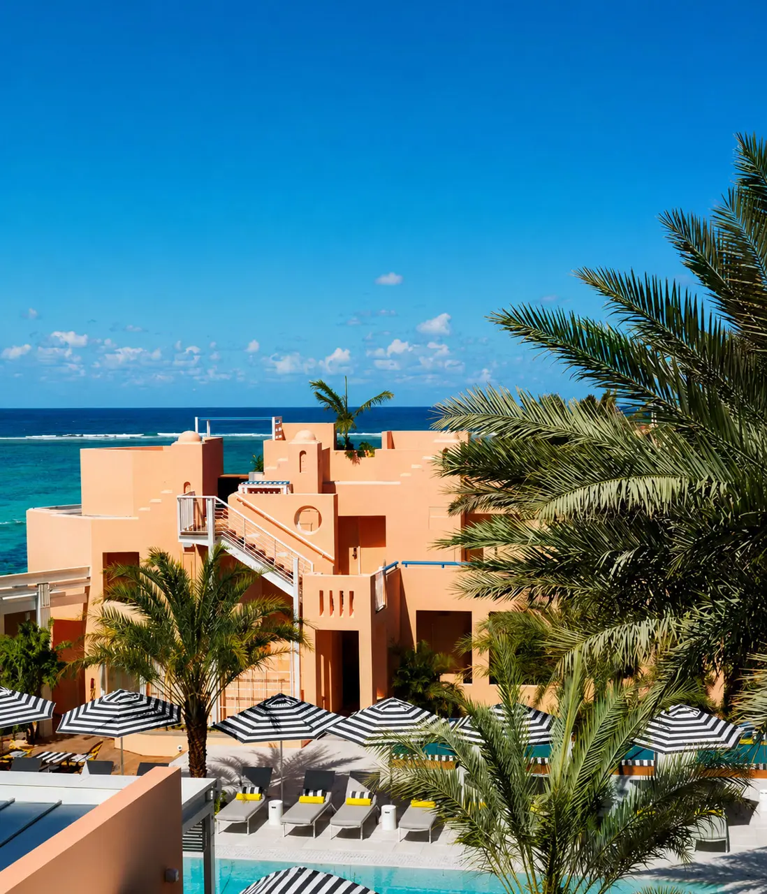 Tropical beachfront resort with terracotta architecture, palm trees, poolside loungers, and ocean views.